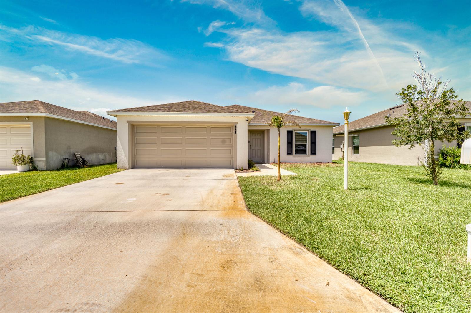 A single-story suburban house with a tan garage, concrete driveway, and a small palm tree — 773 SE 25TH Drive, Okeechobee FL 34974, OKEECHOBEE, FL