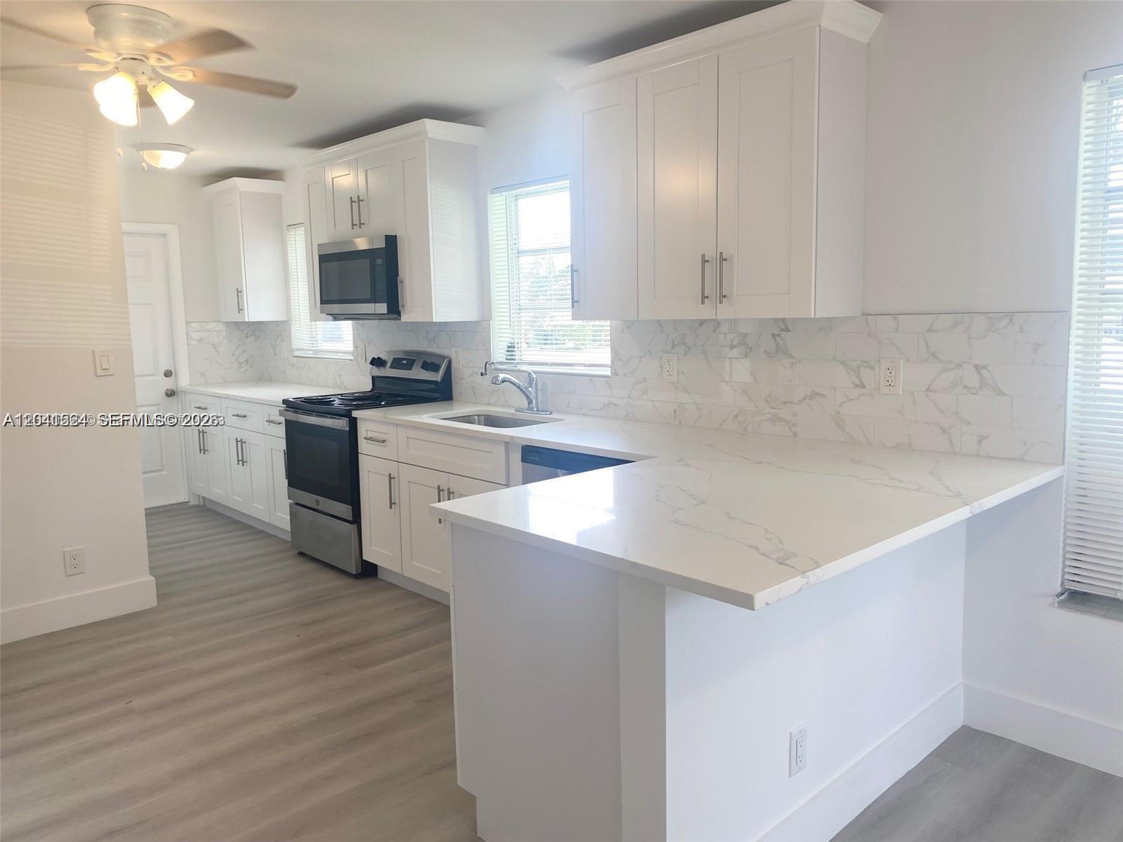 A modern kitchen features white cabinets, marble-style backsplash, stainless steel appliances, and light flooring — 1005 NE 2nd St # 1, Hallandale Beach FL 33009, HALLANDALE BEACH, FL