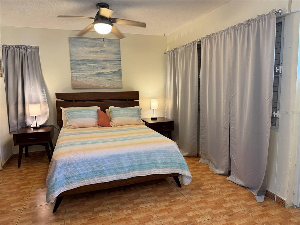 A bedroom features a wooden bed frame, striped bedding, two nightstands, and light gray curtains — COND. LAS CABANAS CARR 466 KM 8.9 UNIT D2, ISABELA, PR