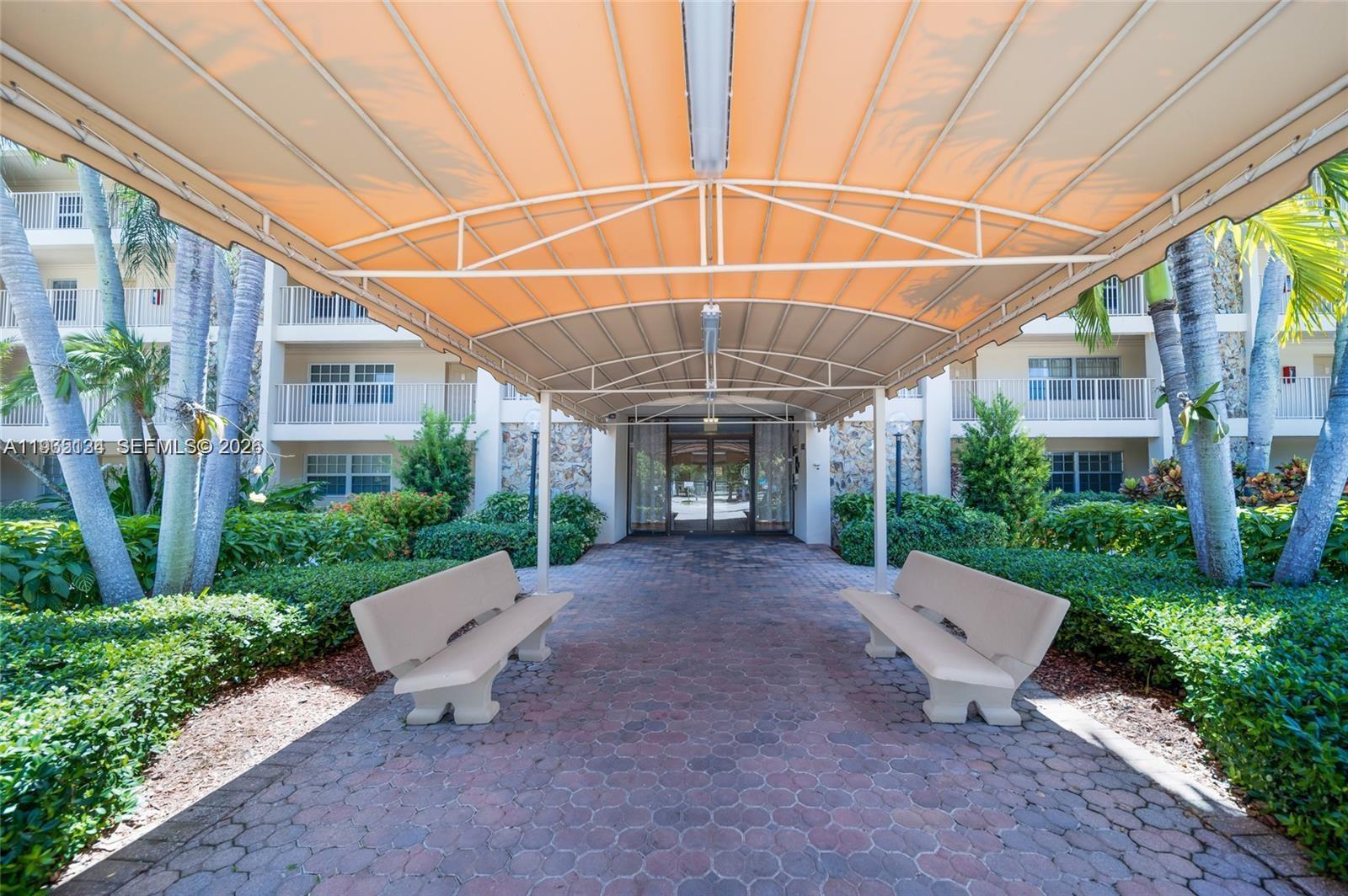 A covered walkway with two benches leads to the glass entrance of a residential building — 4030 W Palm Aire Dr # 104, Pompano Beach FL 33069, POMPANO BEACH, FL
