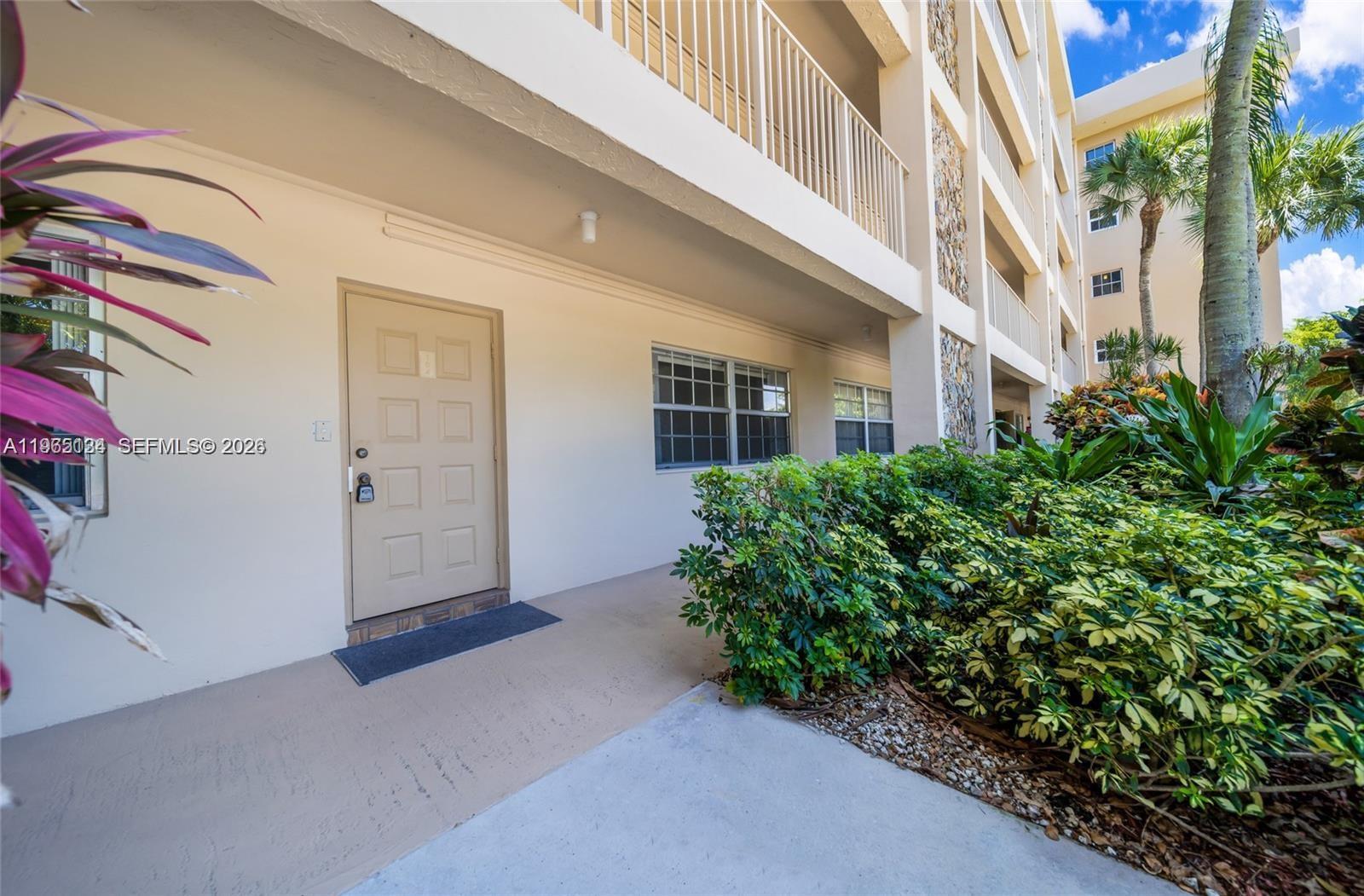 The exterior of a multi-story building features a ground-level entrance door and landscaped bushes — 4030 W Palm Aire Dr # 104, Pompano Beach FL 33069, POMPANO BEACH, FL