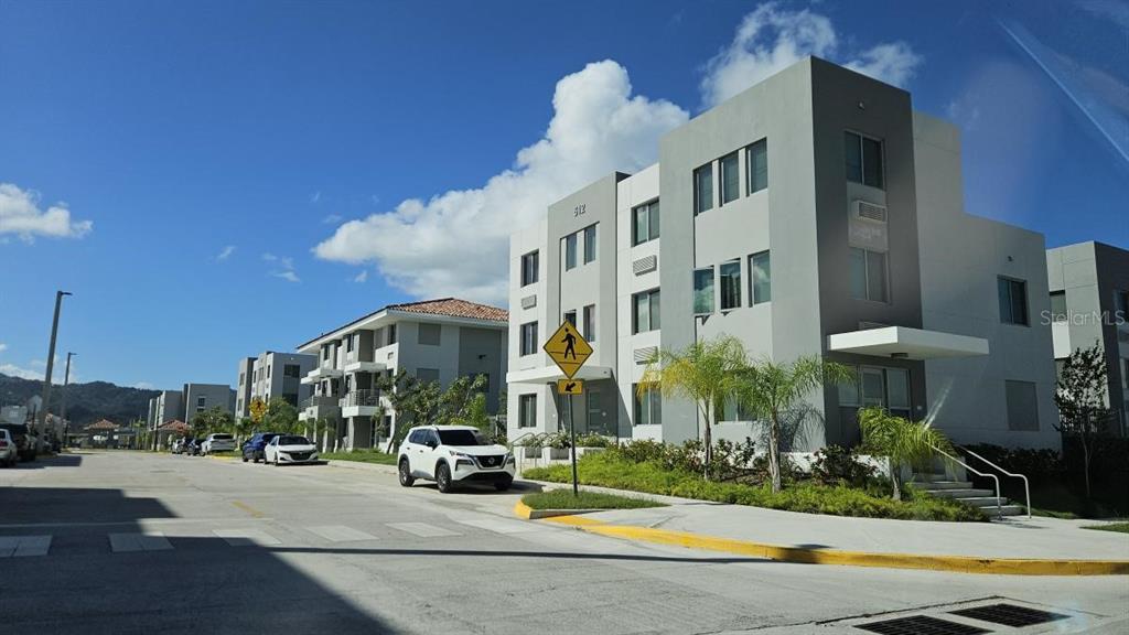 A modern multi-story apartment building stands along a paved street with a white SUV parked — PR-1 INT. SAN CARLOS ST., CAGUAS, PR