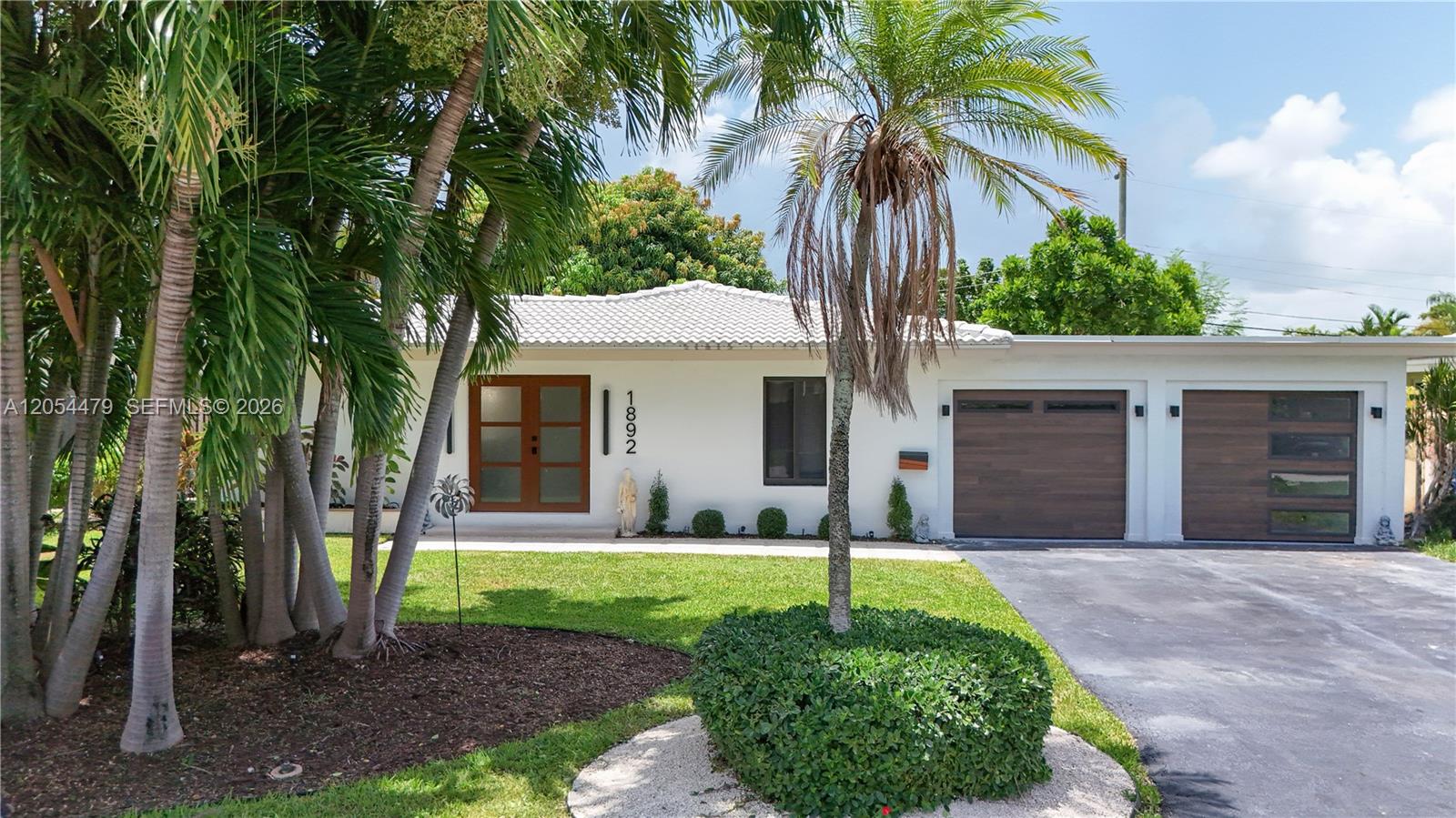 A white single-story house features a double garage, wooden doors, and a palm-filled front yard — 1892 NE 33rd St, Oakland Park FL 33306, OAKLAND PARK, FL
