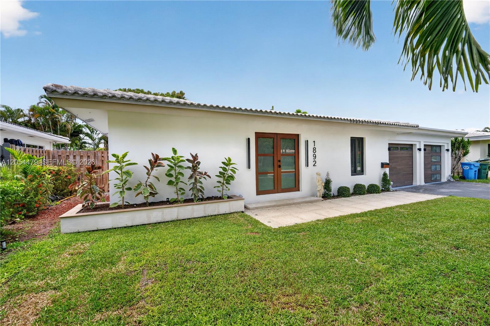 The exterior of a white, single-story house features a front garden bed and double doors — 1892 NE 33rd St, Oakland Park FL 33306, OAKLAND PARK, FL