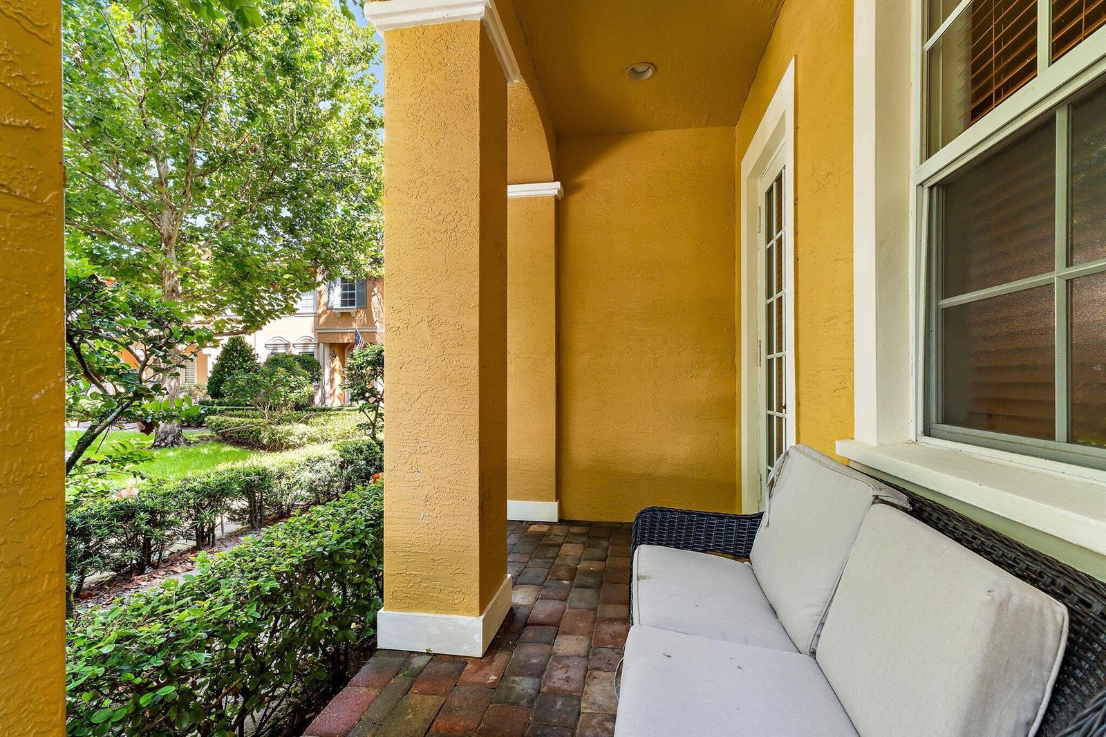 A covered porch with yellow stucco walls, a brick floor, and a wicker outdoor sofa — 150 Lismore Lane, Jupiter FL 33458, JUPITER, FL