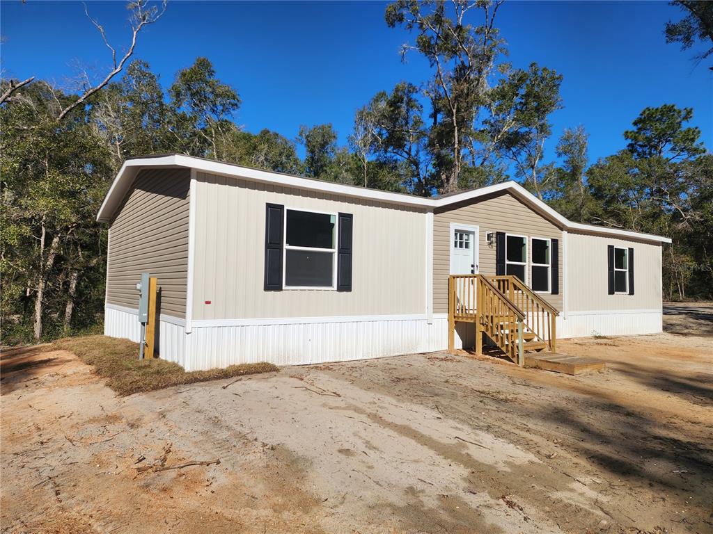 A tan manufactured home with black shutters sits on a grassy lot near trees — 7513 W DUNKLIN STREET, DUNNELLON, FL