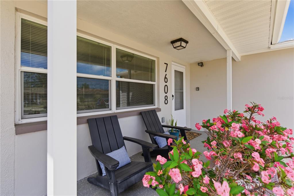 A covered front porch features two black chairs, a white door, and pink flowering plants — 7608 SUE ELLEN DRIVE, PORT RICHEY, FL