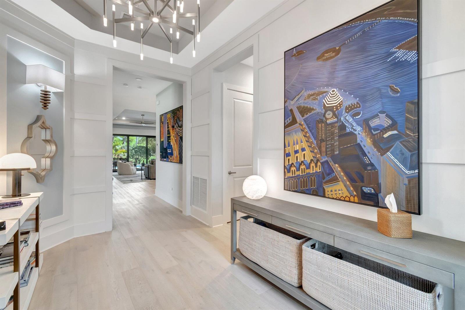 A bright, modern hallway features light wood floors, a large cityscape painting, and a chandelier — 15808 Amelia Terrace, Westlake FL 33470, WESTLAKE, FL