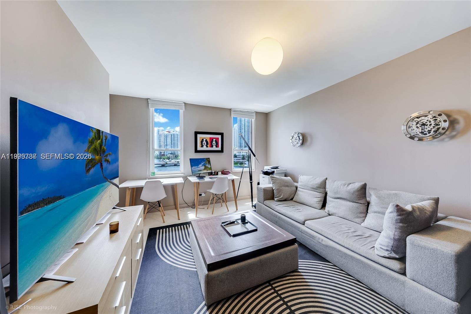 A living room features a grey sofa, a coffee table, two desks, and a television — 701 Brickell Key Blvd # 503, Miami FL 33131, MIAMI, FL