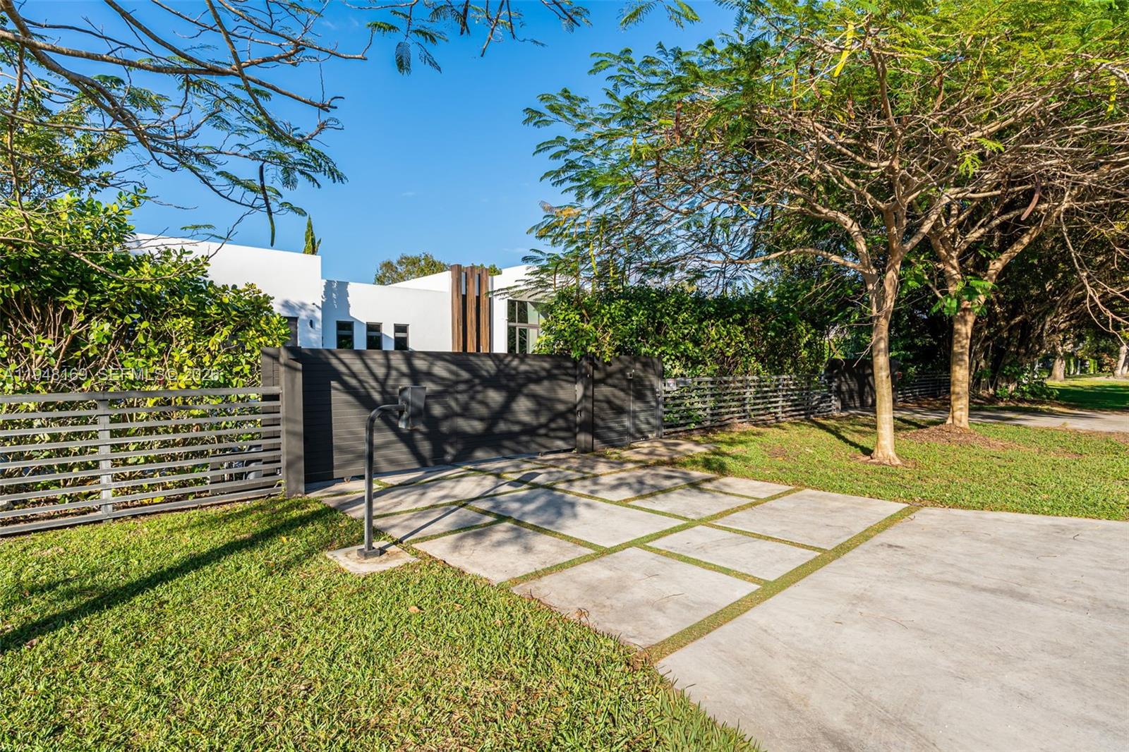 A modern white house is partially obscured by trees behind a dark horizontal slat fence — 6175 SW 128th Street, Pinecrest FL 33156, PINECREST, FL
