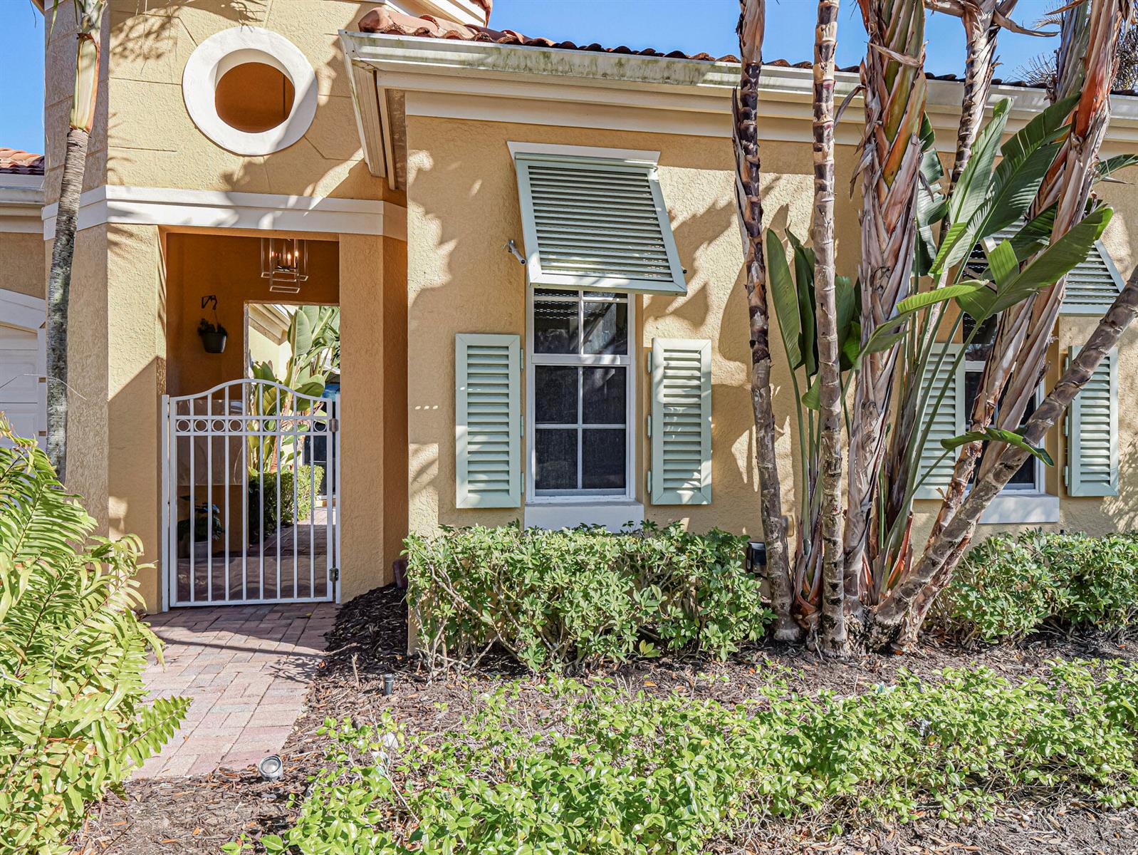 A tan stucco house exterior features a white metal gate, shuttered windows, and landscaping — 9630 E Maiden Court, Vero Beach FL 32963, VERO BEACH, FL