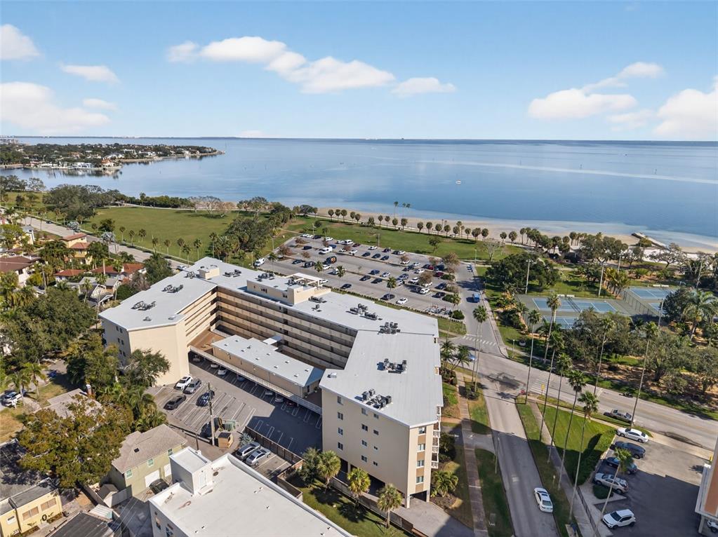 An aerial view shows a multi-story apartment building with a parking lot near a bay — 1200 N SHORE DRIVE NE UNIT 201 furnished, ST PETERSBURG, FL