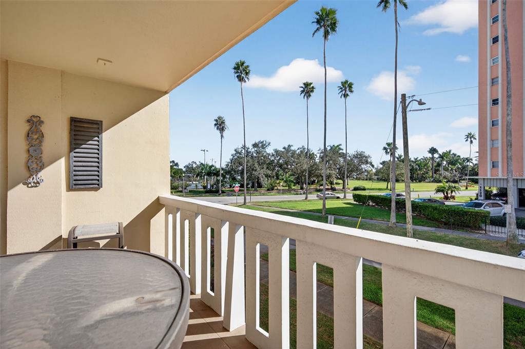 A balcony with a glass-topped table overlooks a street, palm trees, and a neighboring building — 1200 N SHORE DRIVE NE UNIT 201 furnished, ST PETERSBURG, FL
