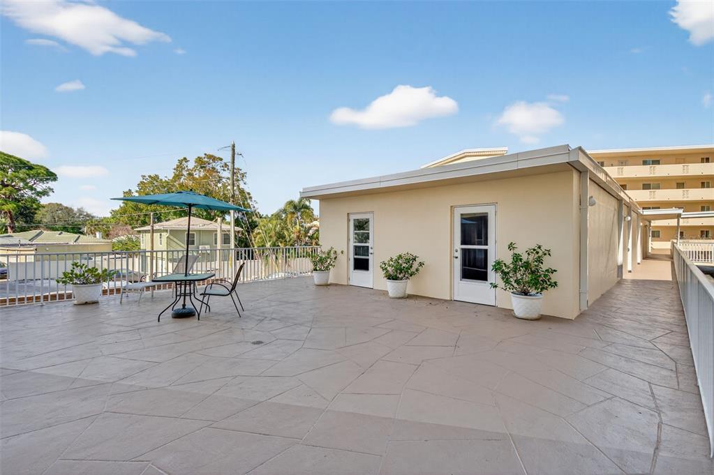 A spacious outdoor rooftop patio features a table, umbrella, potted plants, and two doorways — 1200 N SHORE DRIVE NE UNIT 201 furnished, ST PETERSBURG, FL