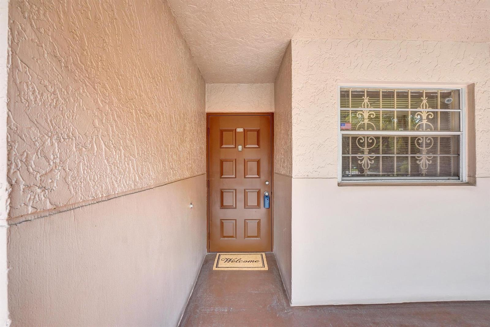 A brown front door with a welcome mat sits in a stucco-walled entryway — 1811 Sabal Palm Drive # 203, Davie FL 33324, DAVIE, FL