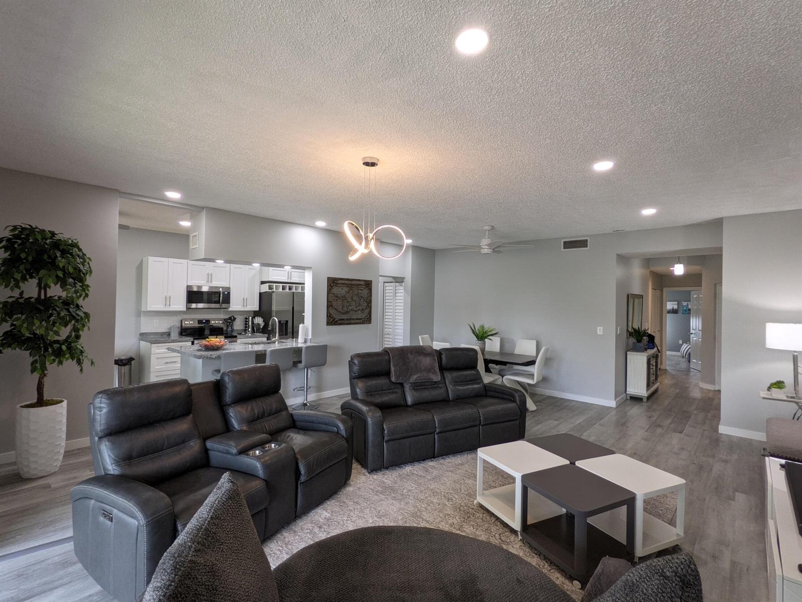 An open-concept living room features gray leather recliners, a modern light fixture, and a kitchen — 105 Half Moon Circle # F2, Hypoluxo FL 33462, HYPOLUXO, FL