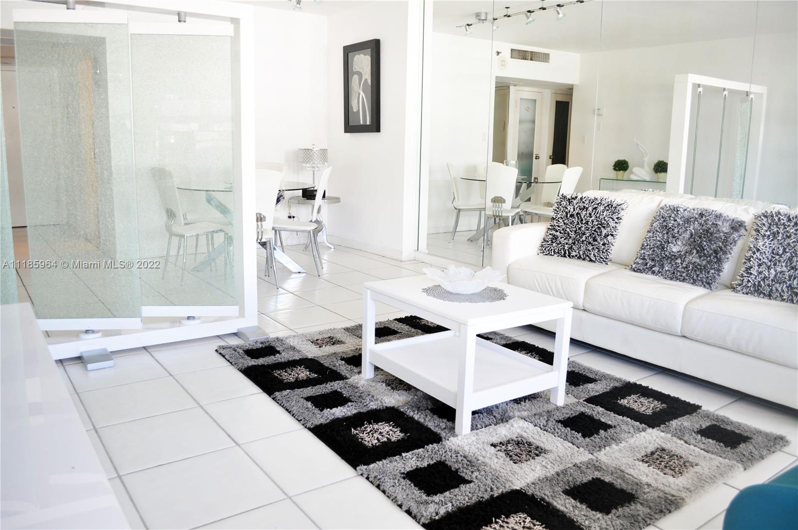 A living room with a white sofa, patterned rug, and a glass room divider — 100 Lincoln Rd # 910, Miami Beach FL 33139, MIAMI BEACH, FL