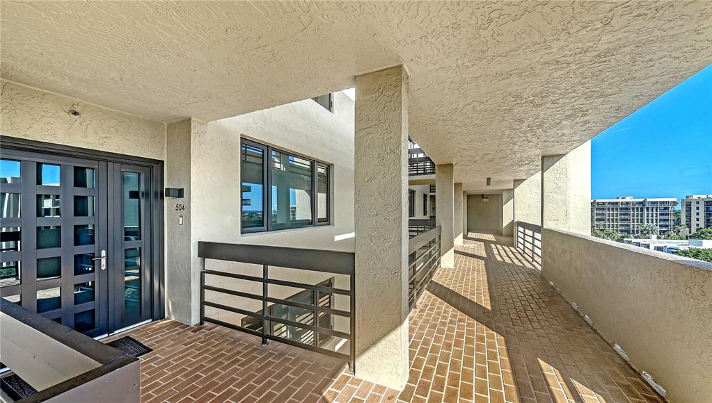 An outdoor balcony walkway features a dark gray door, brick flooring, and metal railings — 1055 GULF OF MEXICO DRIVE UNIT 504, LONGBOAT KEY, FL