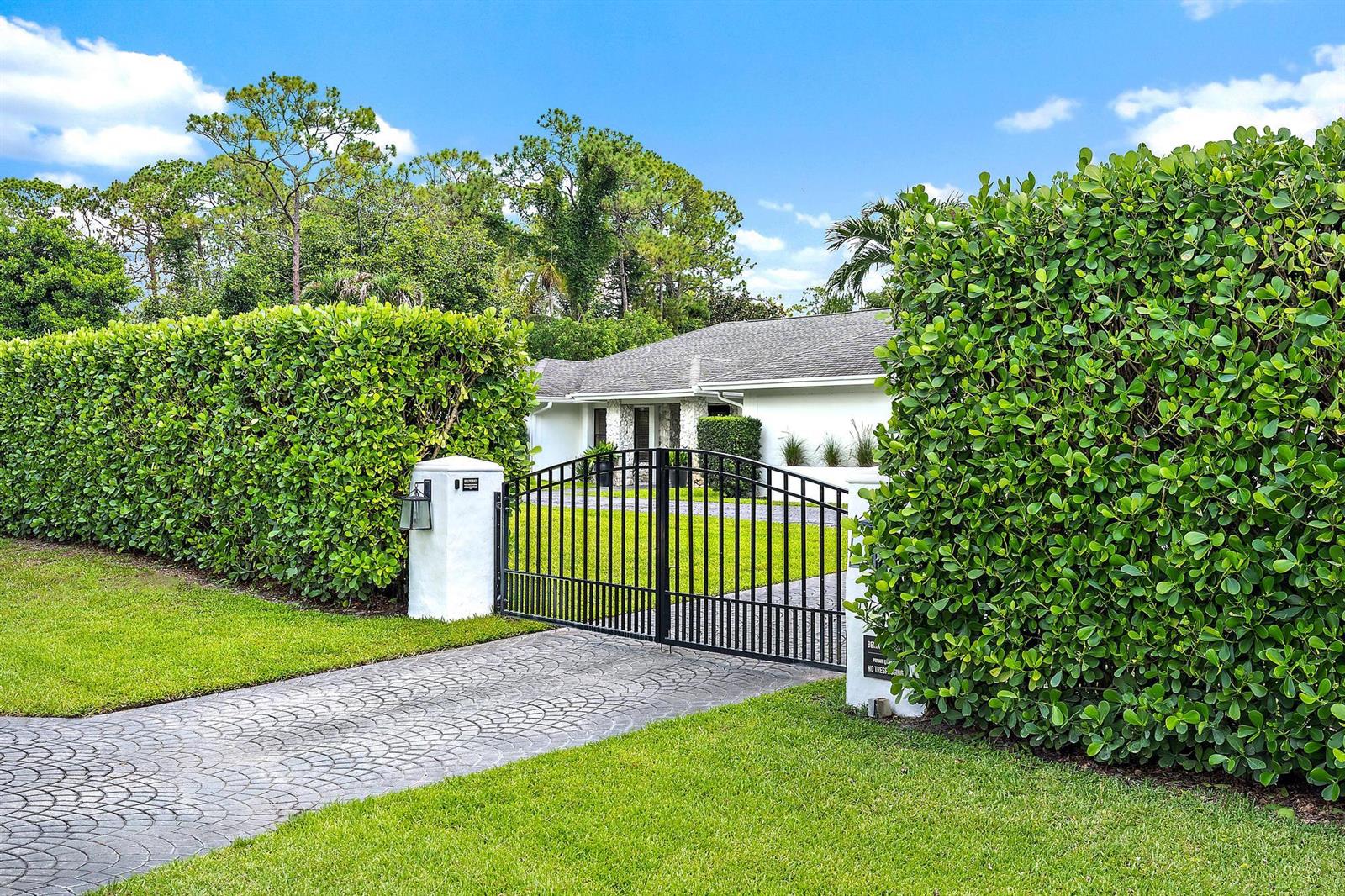 A gated driveway leads to a white house surrounded by tall, manicured green hedges — 14039 Greentree Trail, Wellington FL 33414, WELLINGTON, FL