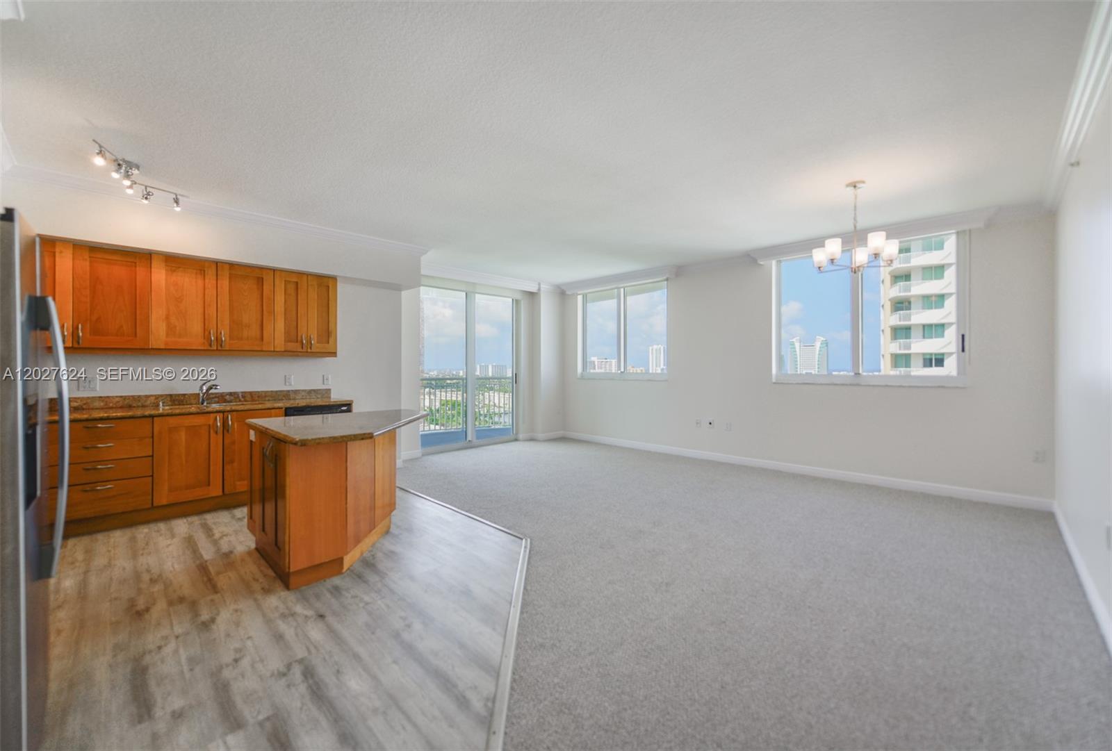 An open-concept kitchen with wood cabinets and a peninsula overlooks a carpeted living area — 1745 E Hallandale Beach Blvd # 1903W, Hallandale Beach FL 33009, HALLANDALE BEACH, FL