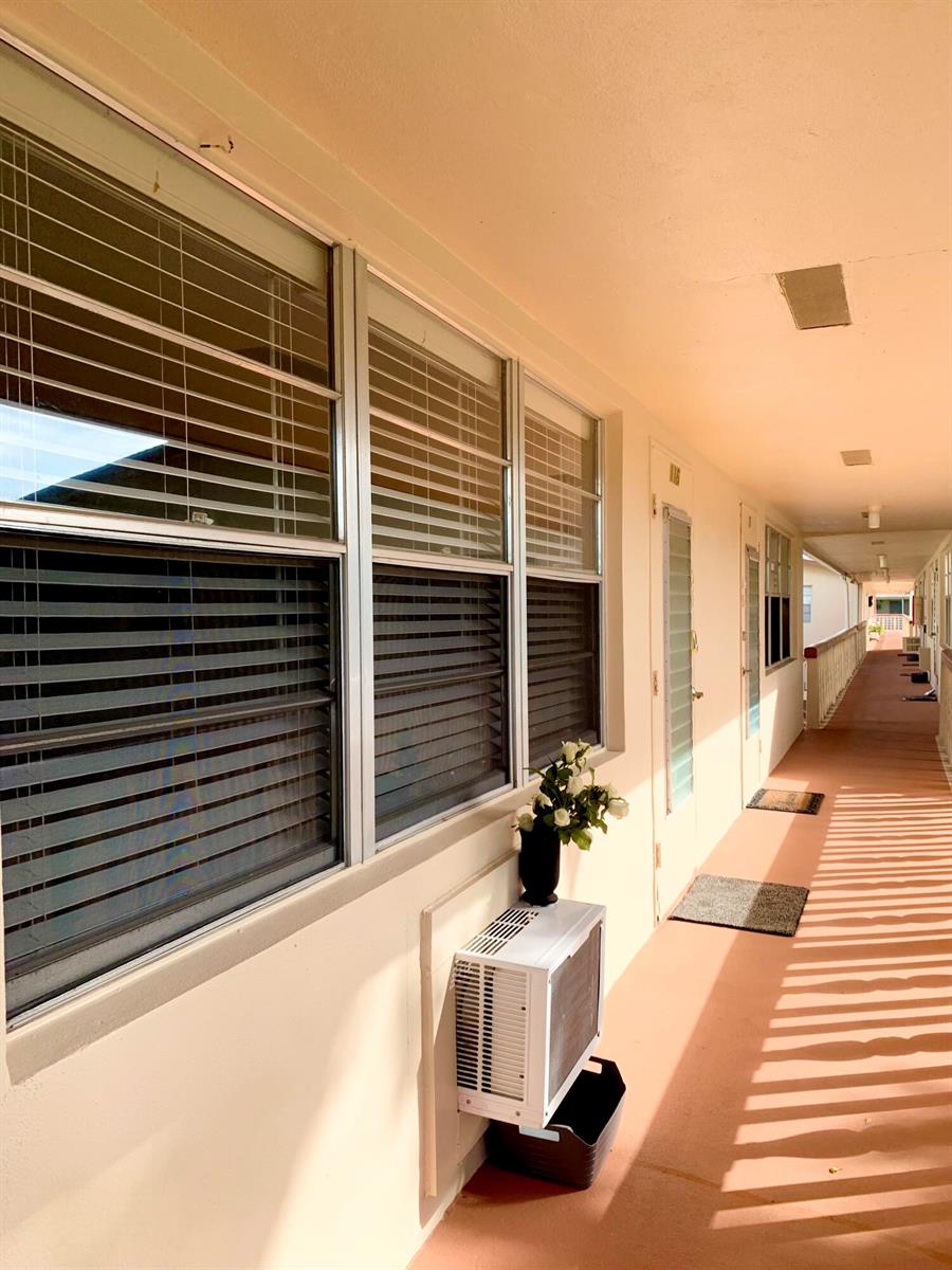 An exterior walkway of an apartment building features windows with blinds and a window air-conditioner — 116 Canterbury E, West Palm Beach FL 33417, WEST PALM BEACH, FL