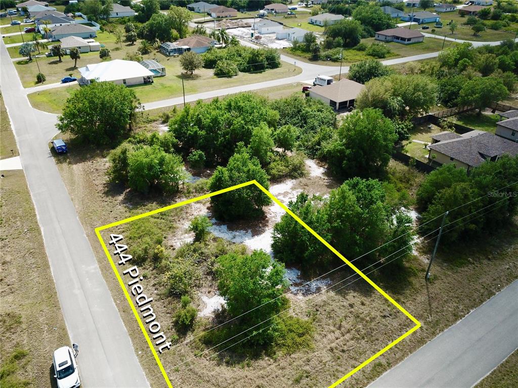 An aerial view shows a vacant, tree-covered corner lot outlined in yellow between two roads — 444 PIEDMONT STREET, LEHIGH ACRES, FL