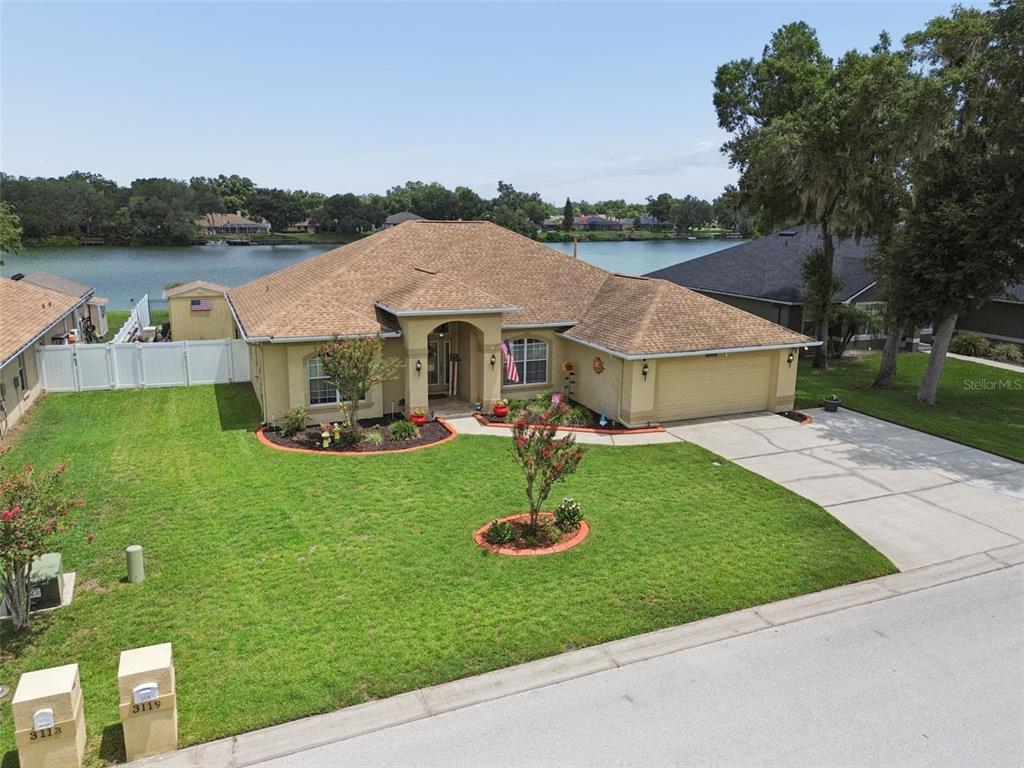 A single-story house with a tan stucco exterior and a brown shingled roof faces a lake — 3119 BLACKWATER OAKS WAY, MULBERRY, FL