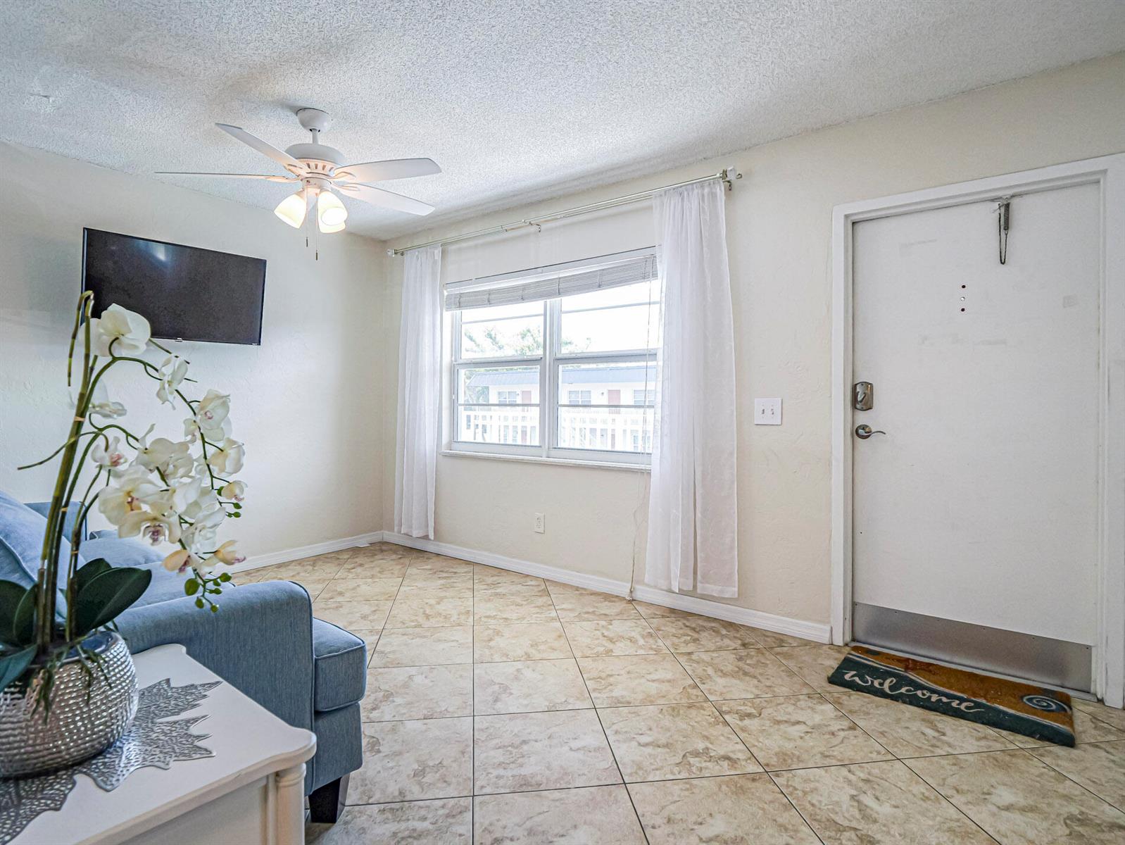 A living room features tile flooring, a ceiling fan, a wall-mounted television, and an entryway — 923 22nd Place # 219, Vero Beach FL 32960, VERO BEACH, FL
