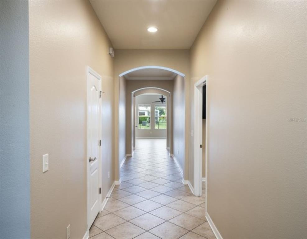 A hallway with beige walls, tile flooring, and arched doorways leading to a bright living area — 7627 STONEY HILL DRIVE, WESLEY CHAPEL, FL