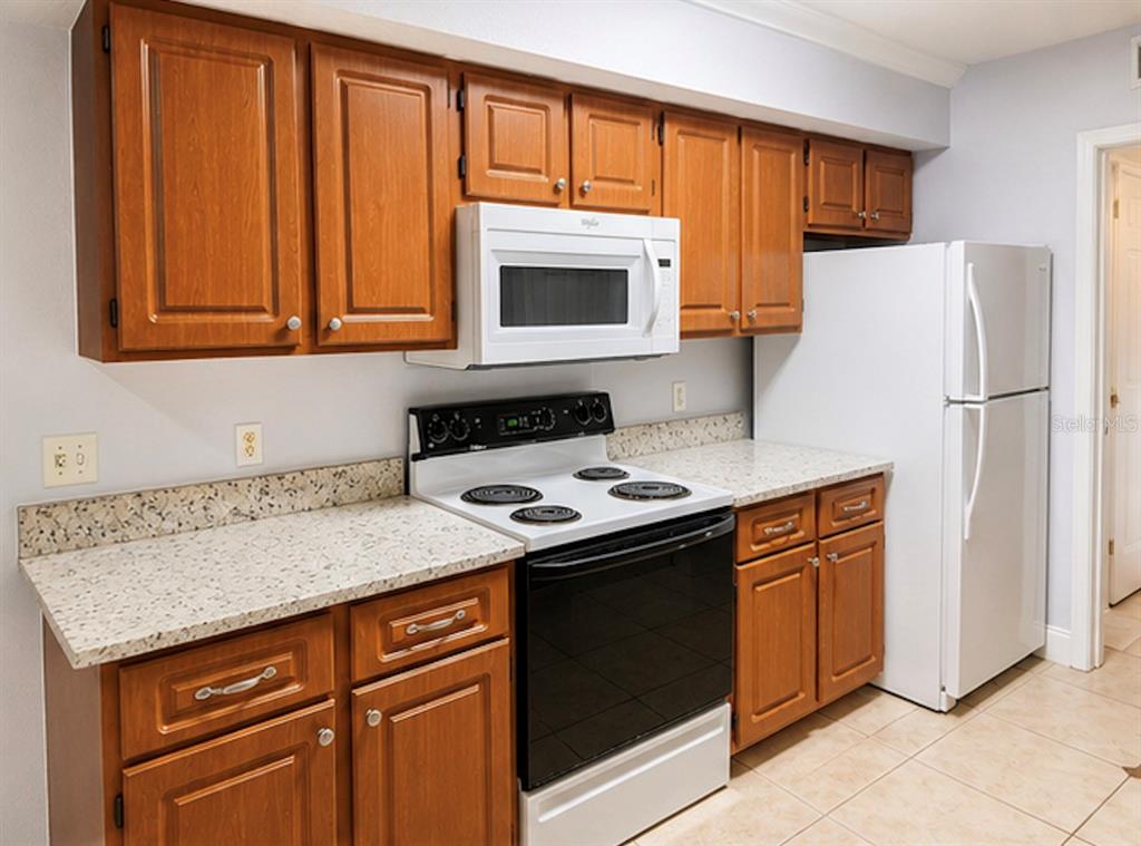 A kitchen features wood cabinets, white appliances, and speckled countertops over tile flooring — 738 CREST PINES DRIVE UNIT 728, ORLANDO, FL