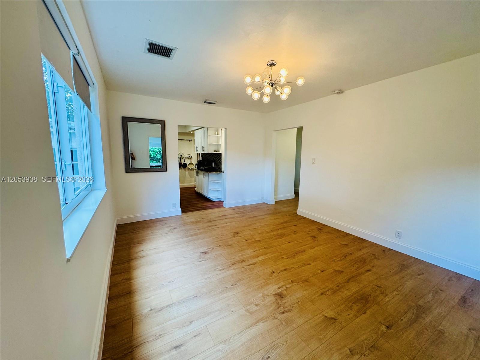 An empty room features light wood flooring, white walls, a modern chandelier, and a kitchen view — 920 N 26th Ave # 0, Hollywood FL 33020, HOLLYWOOD, FL
