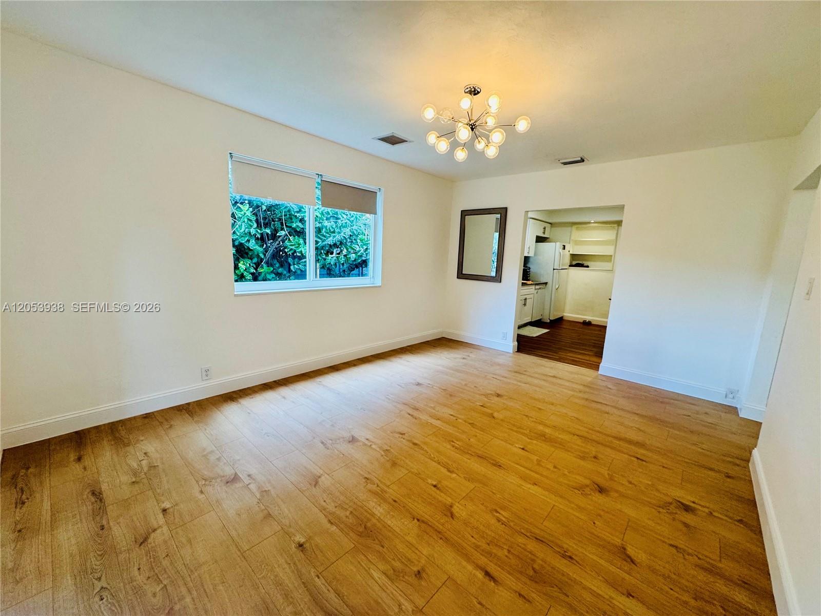 An empty room features light wood-look flooring, a modern chandelier, and a view into a kitchen — 920 N 26th Ave # 0, Hollywood FL 33020, HOLLYWOOD, FL