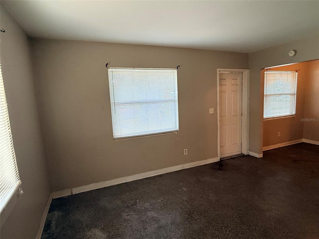 An empty room features beige walls, gray carpeting, two windows with blinds, and a door — 1273 S BETTY LANE, CLEARWATER, FL