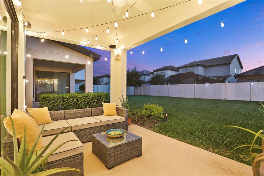 A covered patio features wicker furniture with yellow pillows and string lights overlooking a backyard — 1418 LONE FEATHER TRAIL, WINTER PARK, FL