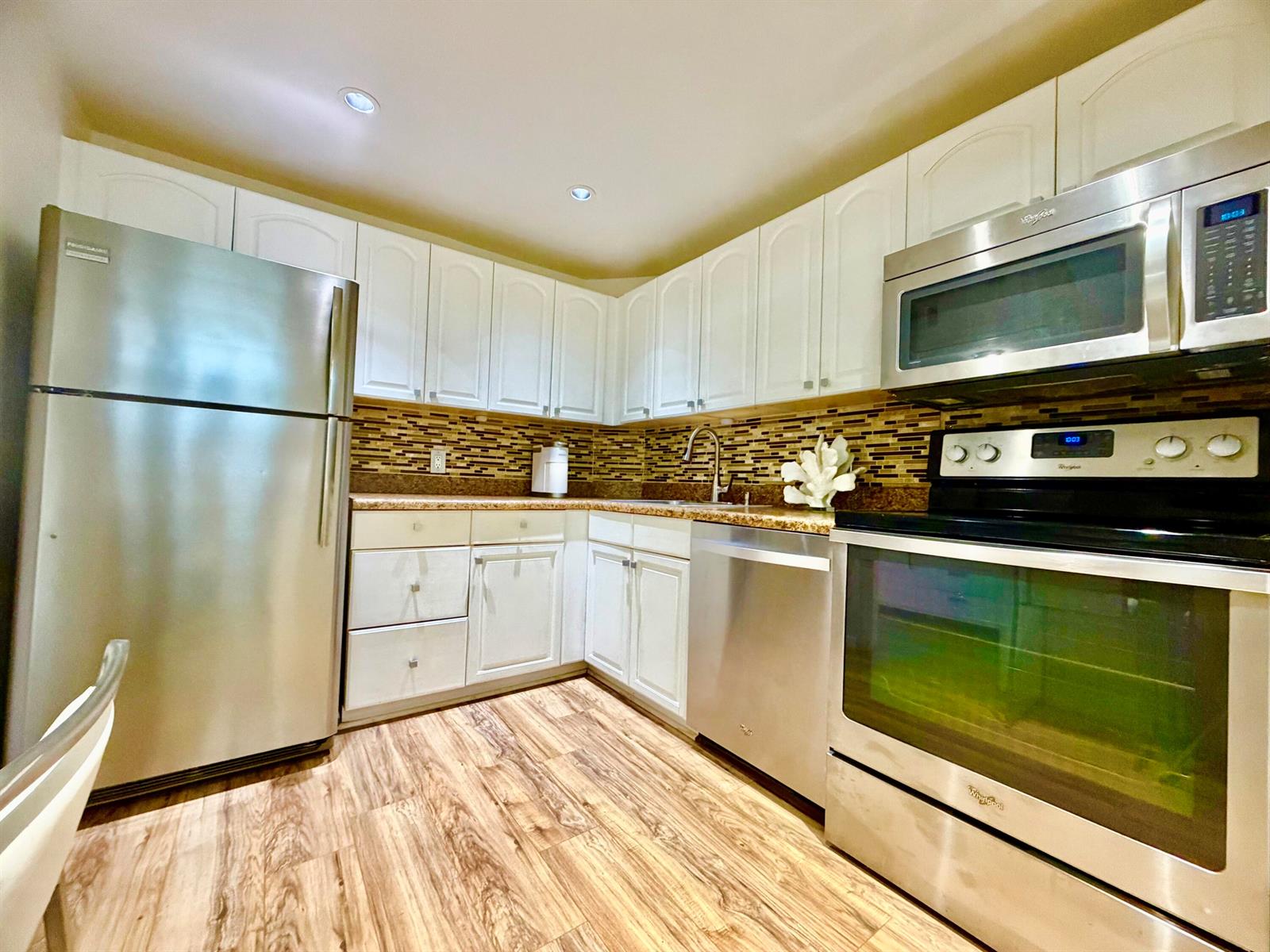 A kitchen features white cabinetry, stainless steel appliances, a tiled backsplash, and wood-look flooring — 3100 N Palm Aire Drive # 706, Pompano Beach FL 33069, POMPANO BEACH, FL