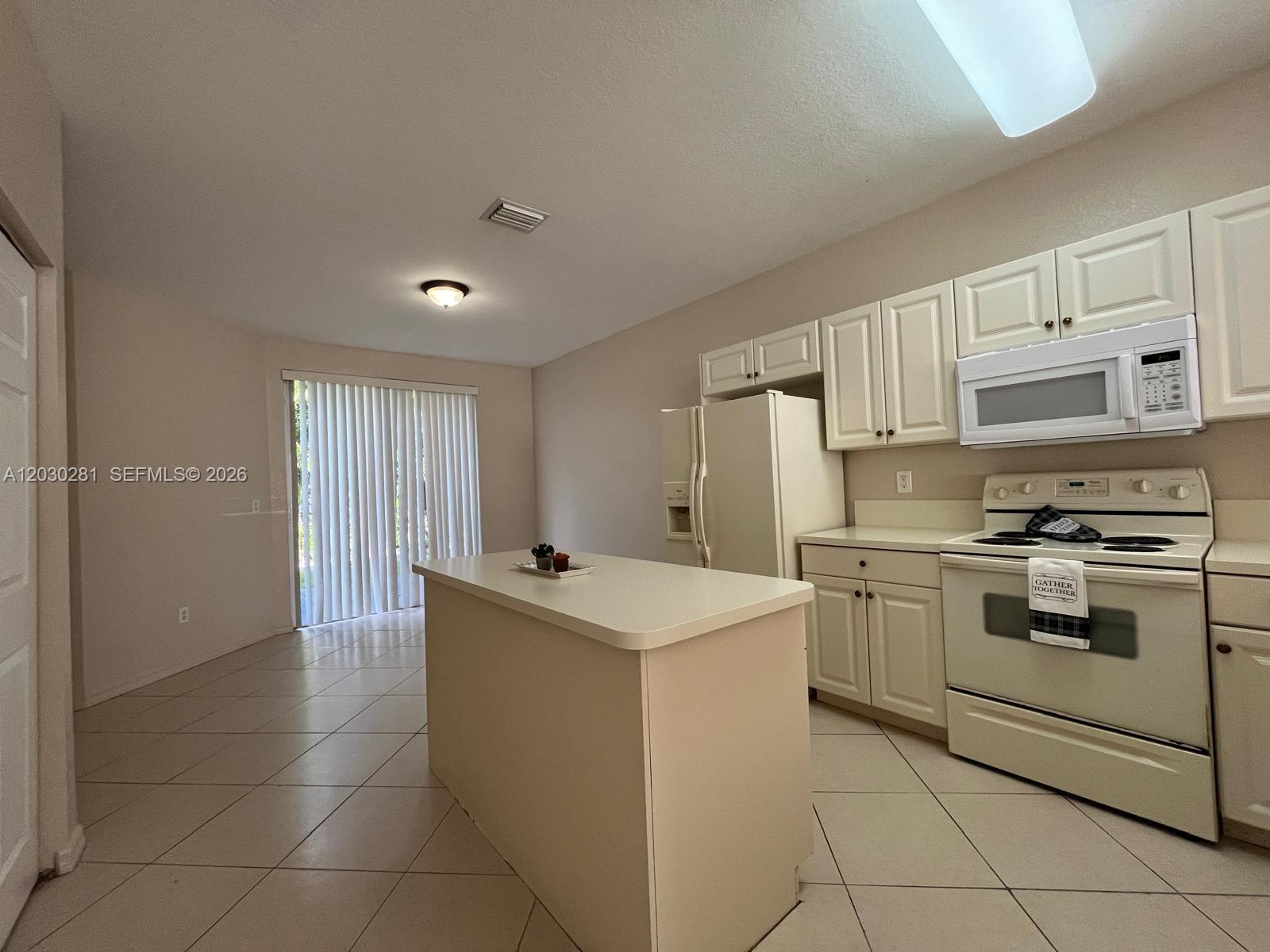 A kitchen features off-white cabinets, a central island, and tile flooring near a sliding door — 1932 Alamanda Way # 1932, Riviera Beach FL 33404, RIVIERA BEACH, FL