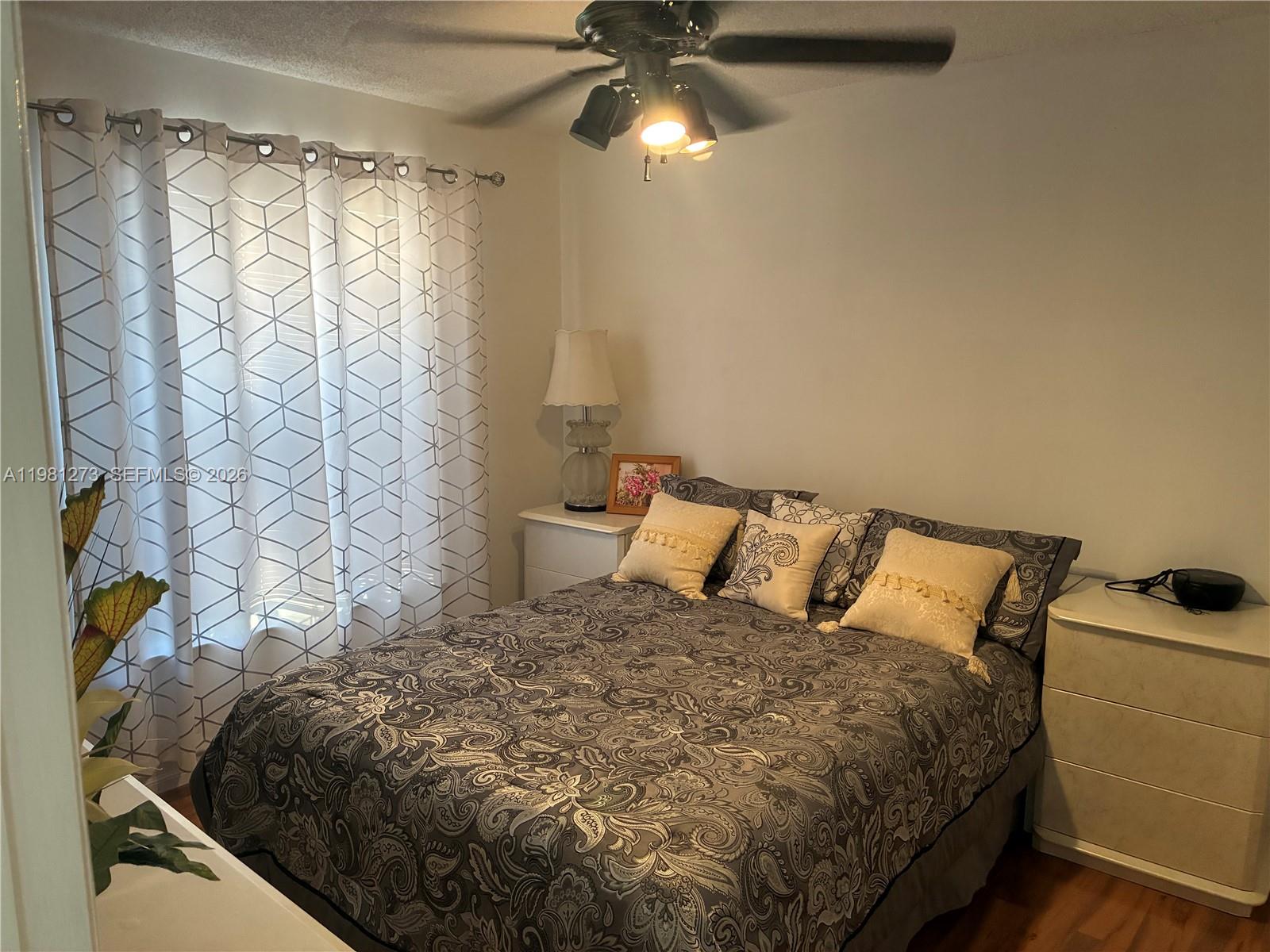 A bedroom features a bed with patterned bedding, two white nightstands, and a ceiling fan — 18175 NW 61st Ave # 0, Miami FL 33015, MIAMI, FL