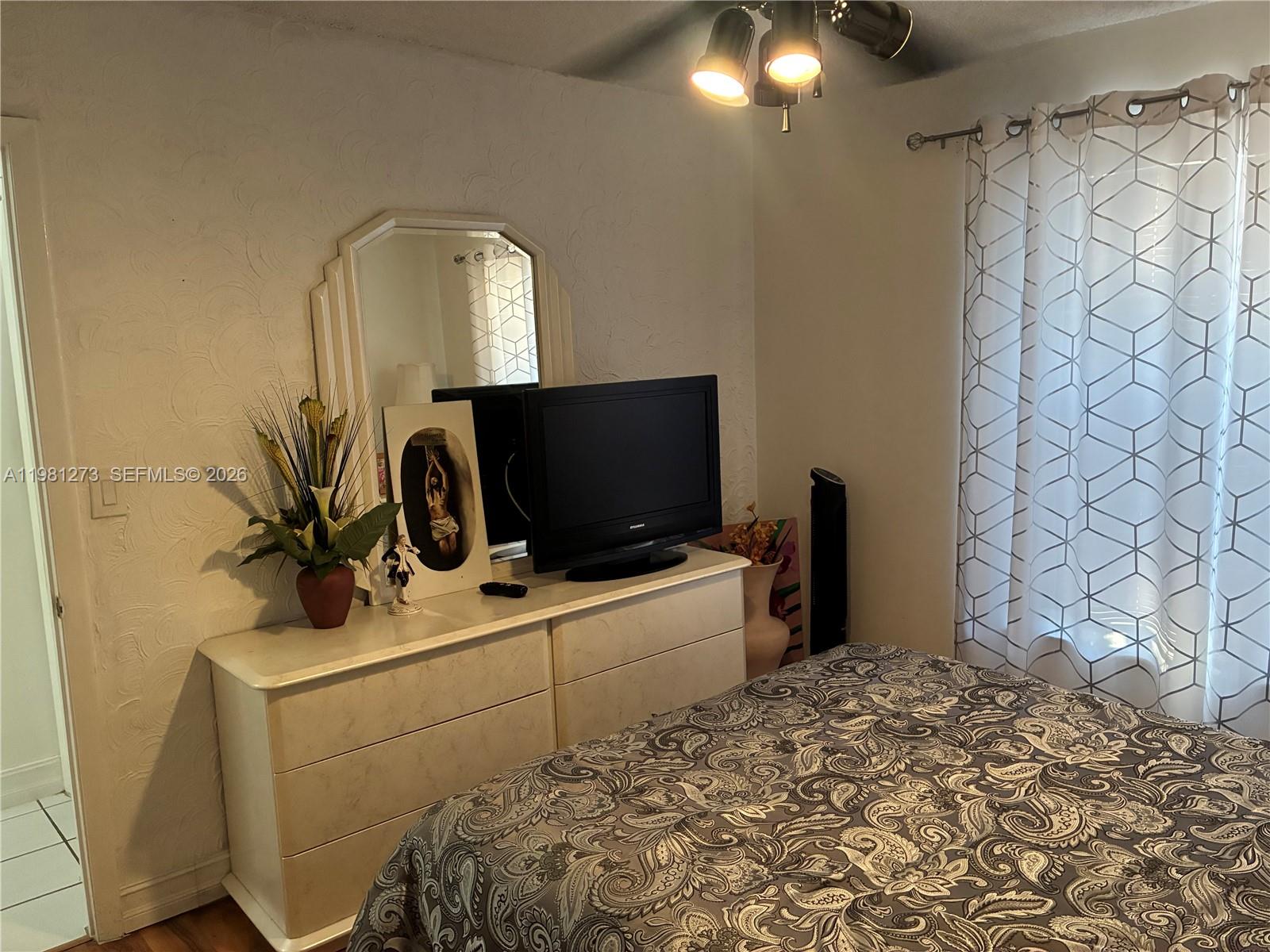 A bedroom features a white dresser with a mirror, a television, and a patterned bedspread — 18175 NW 61st Ave # 0, Miami FL 33015, MIAMI, FL