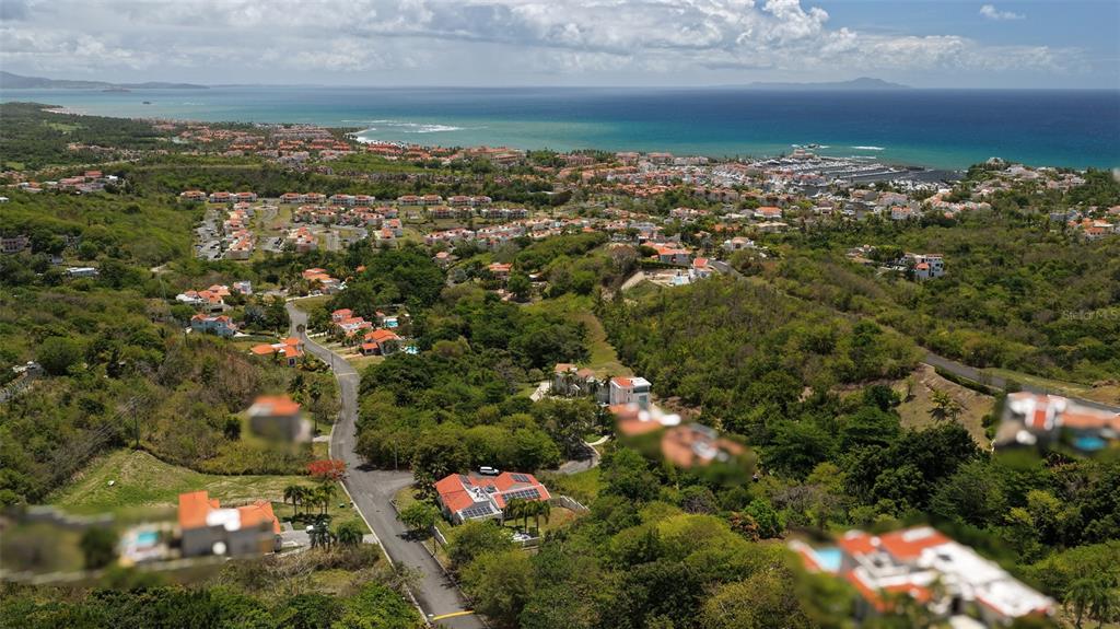 An aerial view shows a coastal residential neighborhood with houses, lush vegetation, and the ocean — 76 HARBOUR LIGHTS DR, HUMACAO, PR