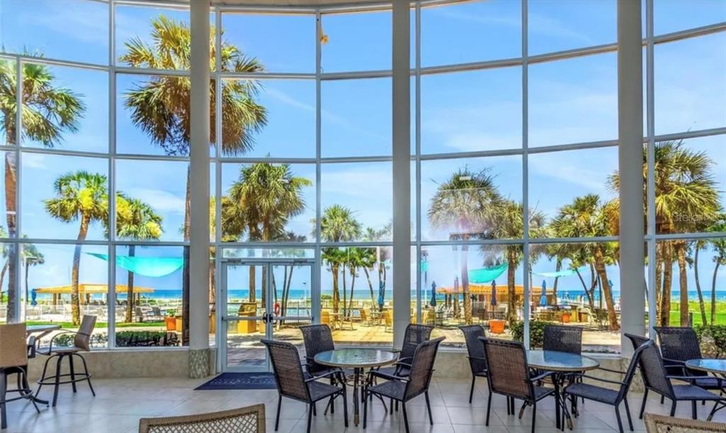 A sunlit indoor seating area features floor-to-ceiling windows overlooking a beach with palm trees — 880 MANDALAY AVENUE UNIT C410, CLEARWATER, FL