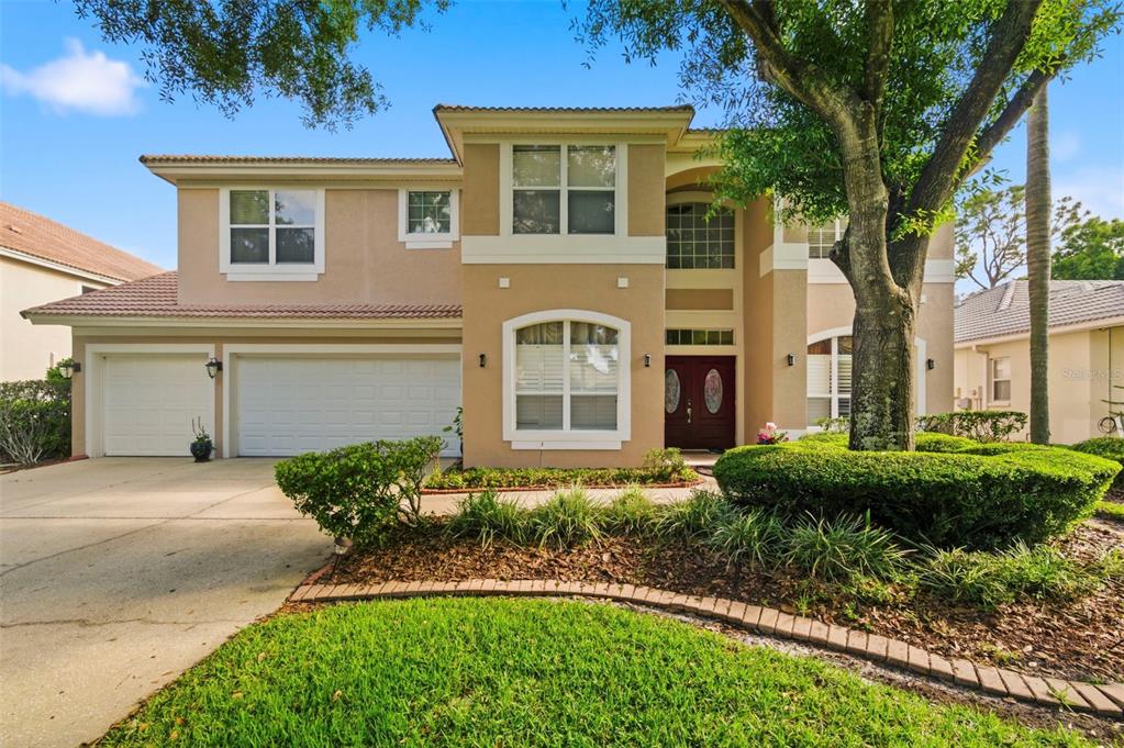 A two-story tan stucco house features a three-car garage and a landscaped front yard — 9207 MEADOW LANE COURT, TAMPA, FL