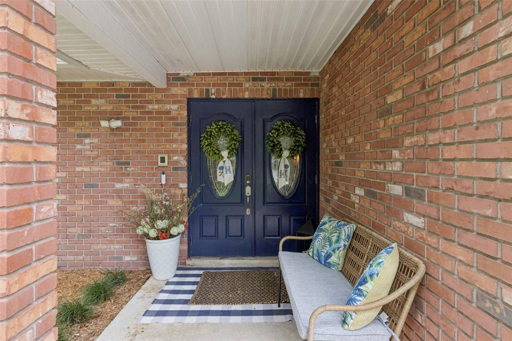A covered brick porch features double navy blue front doors with wreaths and a wicker bench — 204 BIRCH LANE, LAKELAND, FL