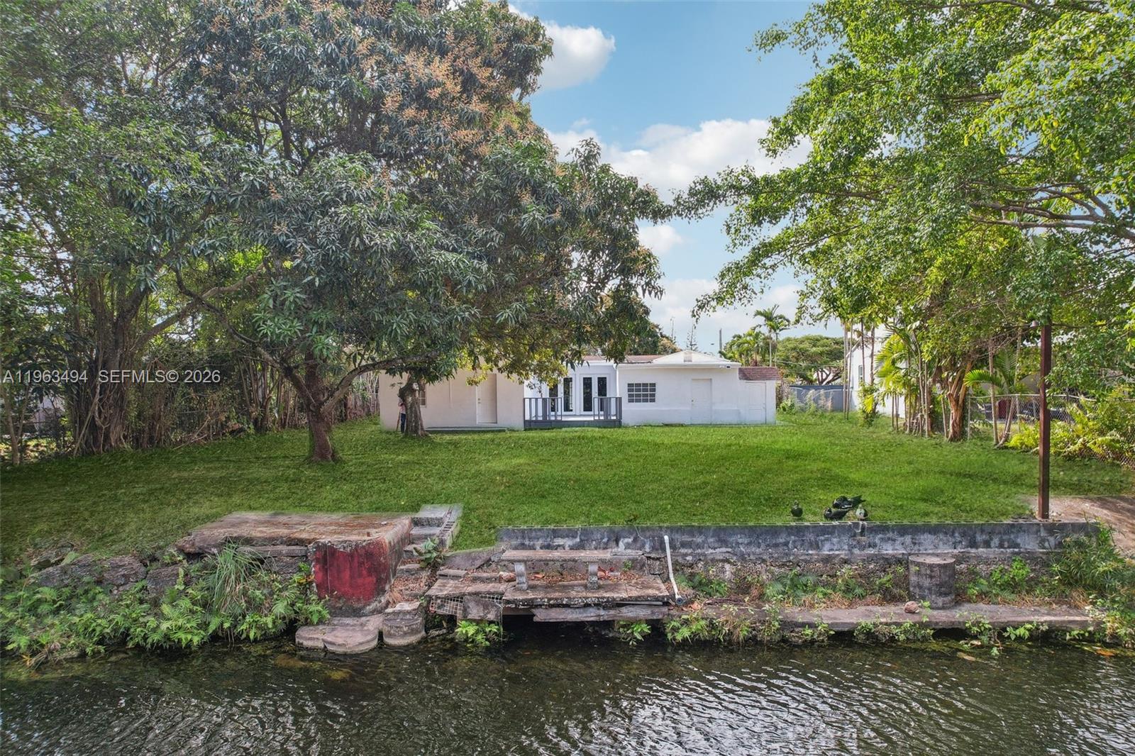 A white house sits on a grassy lot overlooking a canal with a concrete bulkhead — 299 S Biscayne River Dr, Miami FL 33169, MIAMI, FL