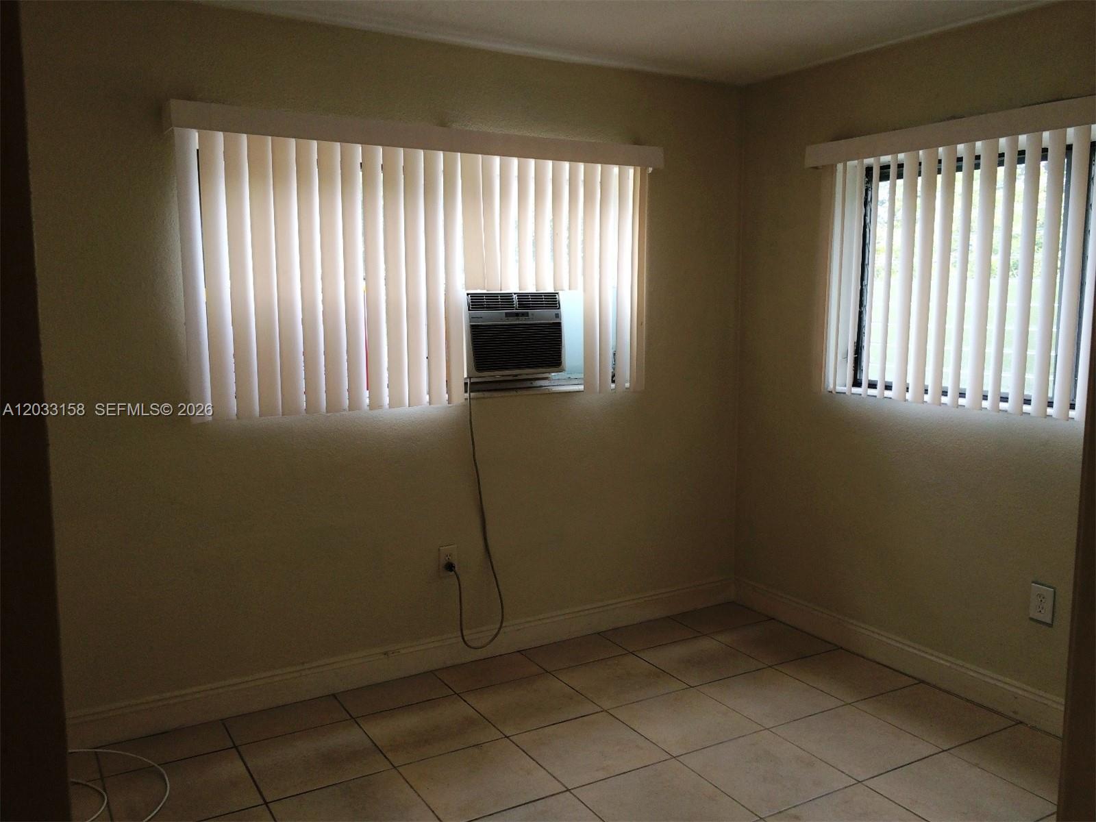 An empty room features beige walls, tiled flooring, two windows with vertical blinds, and an air conditioner — 2441 NW 13 ST # 77, Miami FL 33125, MIAMI, FL