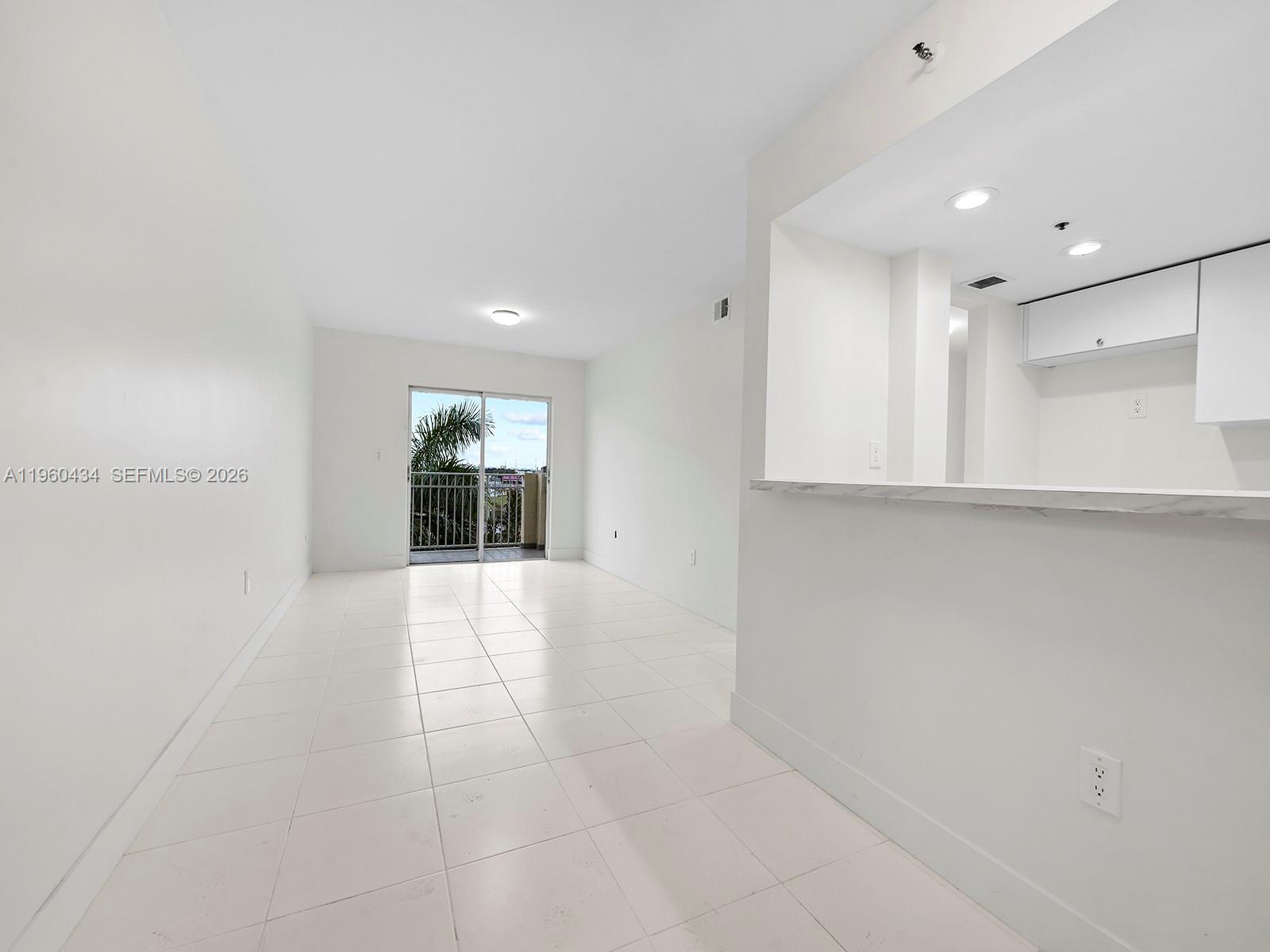 A bright, empty room with white tile floors, a kitchen pass-through, and a balcony door — 1750 NW 27th Ave # 0, Miami FL 33125, MIAMI, FL