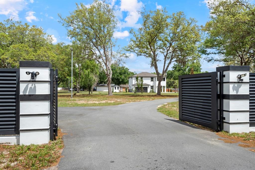 A paved driveway leads through a modern gated entrance toward a two-story white house — 2509 APPY LANE, APOPKA, FL