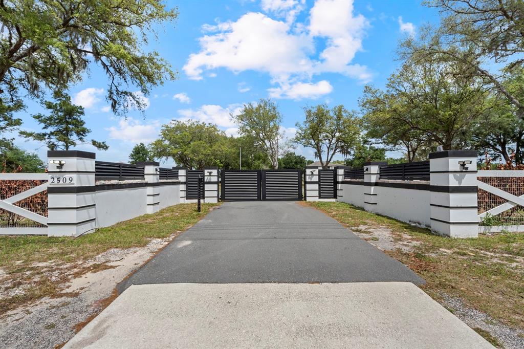A paved driveway leads to a modern black metal gate flanked by white masonry pillars — 2509 APPY LANE, APOPKA, FL