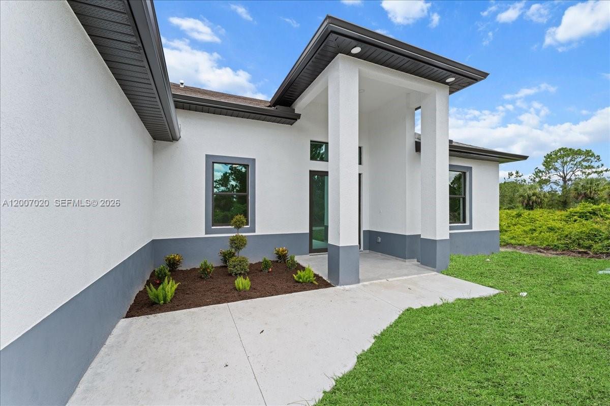 The exterior of a modern house features white stucco walls, gray trim, and a covered entryway — 1033 E Plymouth St E, Lehigh Acres FL 33974, LEHIGH ACRES, FL