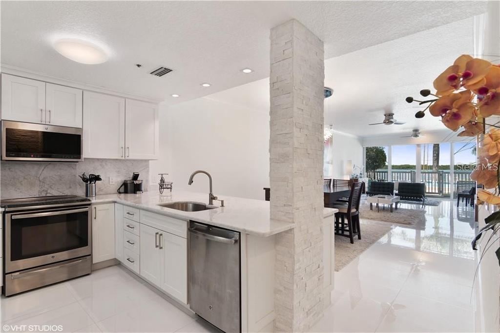 A modern kitchen with white cabinets and stone countertops overlooks a bright, open living area — 615 DREAM ISLAND ROAD UNIT 107, LONGBOAT KEY, FL