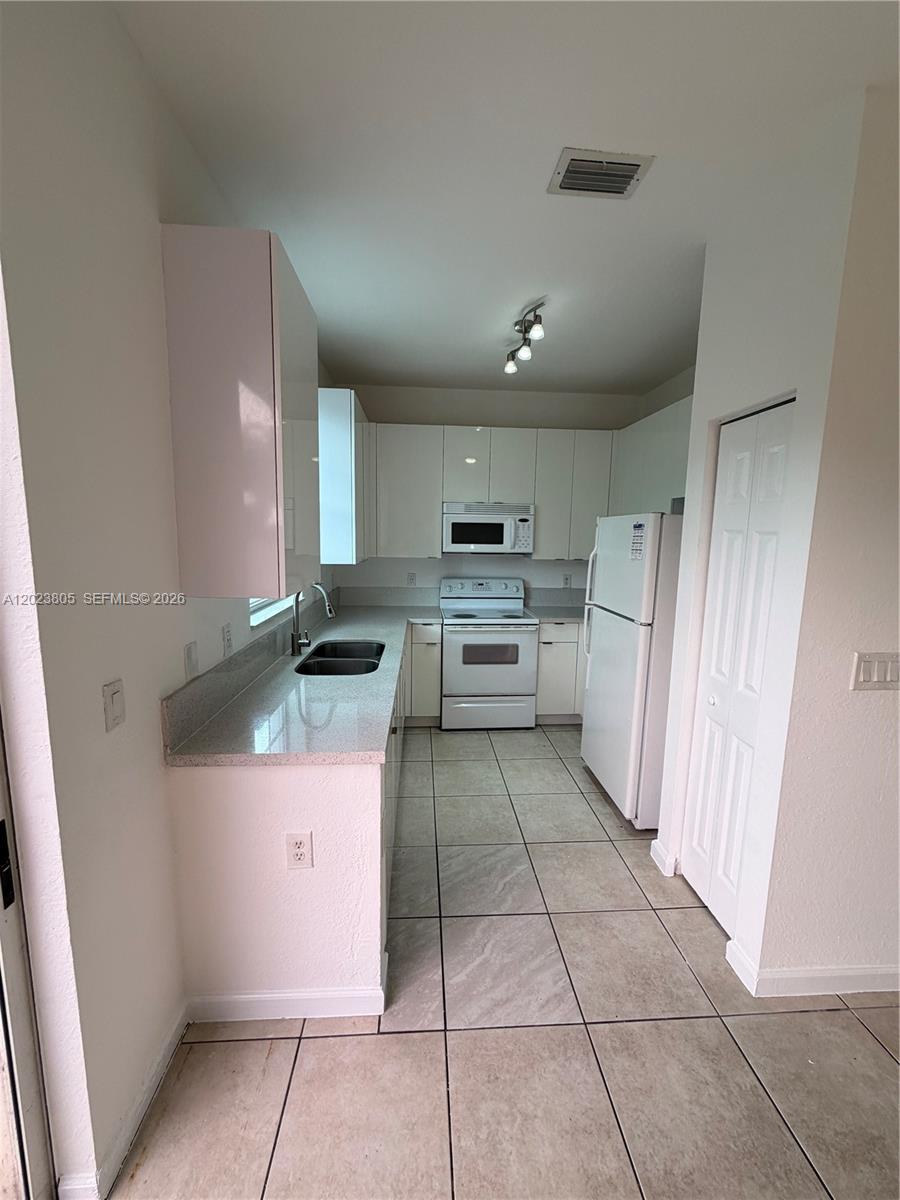 A galley-style kitchen features white cabinets, white appliances, and light-colored tile flooring — 607 NE 21st Ter, Homestead FL 33033, HOMESTEAD, FL